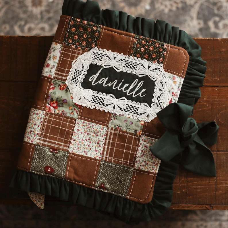 May include: A handmade, quilted journal with a patchwork design in brown, floral, and plaid fabrics. The journal features a white lace-trimmed label with the name "danielle" in cursive. Dark green ruffle trim and a matching bow add a decorative touch.