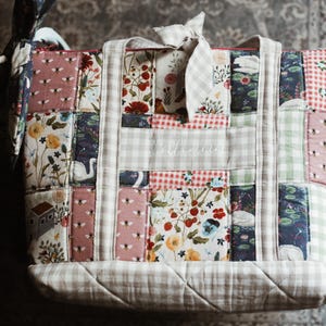 May include: A patchwork tote bag with a variety of floral and patterned fabrics. The bag features a mix of pink, red, and blue floral prints, along with gingham and bee patterns. The bag has a light-colored handle and a bow.