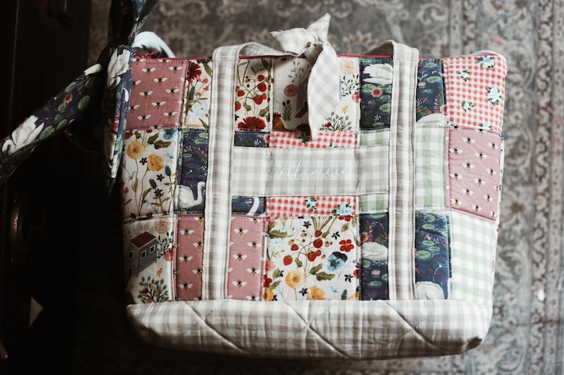 May include: A patchwork tote bag with a variety of floral and patterned fabrics. The bag features a mix of pink, red, and blue floral prints, along with gingham and bee patterns. The bag has a light-colored handle and a bow.