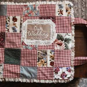 May include: A patchwork wedding album with a red and white gingham design. The album incorporates floral and patterned fabric squares, a lace-trimmed edge, and a handle. The text "Our Wedding 11/16/2024" is embroidered on the front.