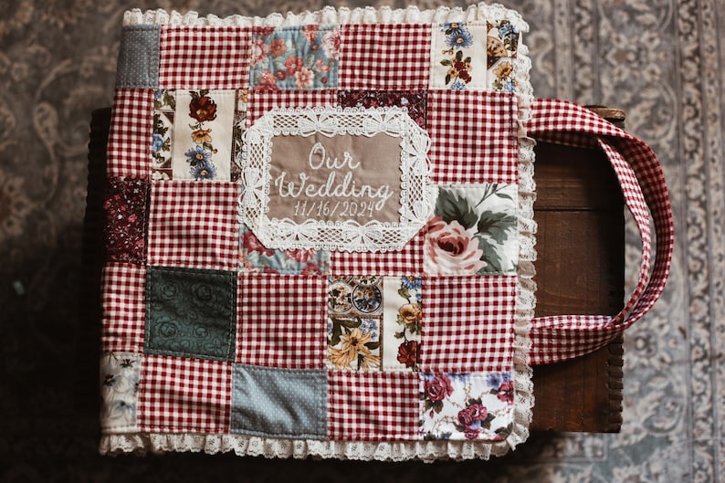 May include: A patchwork wedding album with a red and white gingham design. The album incorporates floral and patterned fabric squares, a lace-trimmed edge, and a handle. The text "Our Wedding 11/16/2024" is embroidered on the front.