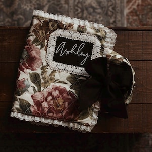 May include: A quilted fabric book cover featuring a floral pattern in shades of burgundy, green, and cream. The cover is trimmed with white lace and includes a black rectangular patch with the name Ashley embroidered in white, plus a black bow.