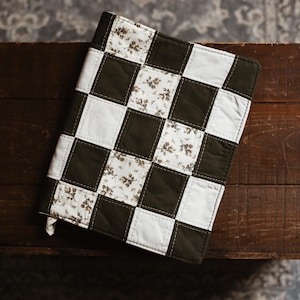 May include: A handmade, quilted book cover with a checkerboard pattern. Alternating squares are white, dark green, and floral print. The cover is resting on a dark wooden surface.