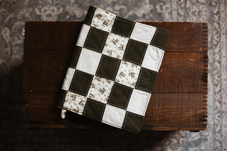 May include: A handmade, quilted book cover with a checkerboard pattern. Alternating squares are white, dark green, and floral print. The cover is resting on a dark wooden surface.