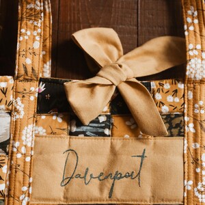 May include: A quilted tote bag with a mustard yellow floral pattern and a bow. The bag features a rectangular patch with the name "Davenport" embroidered in a script font. The bag's design includes a mix of floral and geometric patterns.