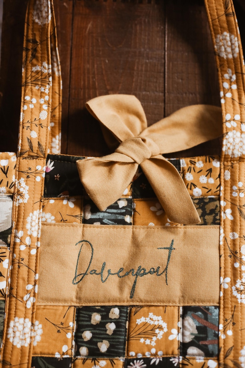 May include: A quilted tote bag with a mustard yellow floral pattern and a bow. The bag features a rectangular patch with the name "Davenport" embroidered in a script font. The bag's design includes a mix of floral and geometric patterns.