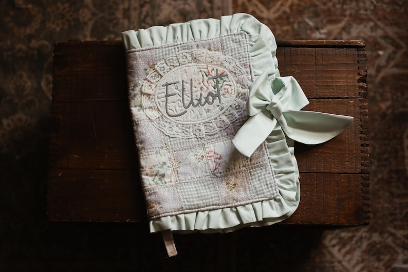 May include: A personalised baby book with the name "Elliot" embroidered on the cover. The book features a patchwork design with floral and gingham patterns in soft pastel colours. A mint green bow secures the book, and the edges are trimmed with a ruffled border.