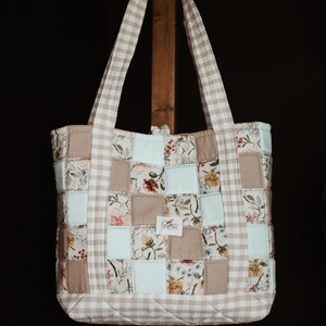 May include: A patchwork tote bag with a beige and white gingham pattern on the handles and trim. The body of the bag is composed of squares in floral prints, solid beige, and light blue. The bag is hanging on a wooden stand.