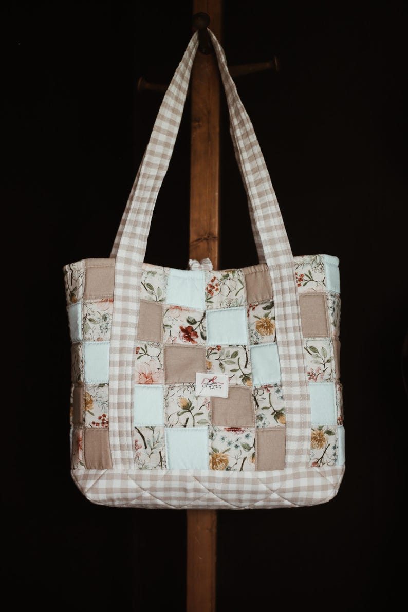 May include: A patchwork tote bag with a beige and white gingham pattern on the handles and trim. The body of the bag is composed of squares in floral prints, solid beige, and light blue. The bag is hanging on a wooden stand.