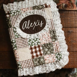 May include: A patchwork journal with a brown oval label embroidered with the name "Alexis" in white script. The journal features a variety of fabric patterns, including gingham, floral, and polka dots, and is trimmed with white lace.