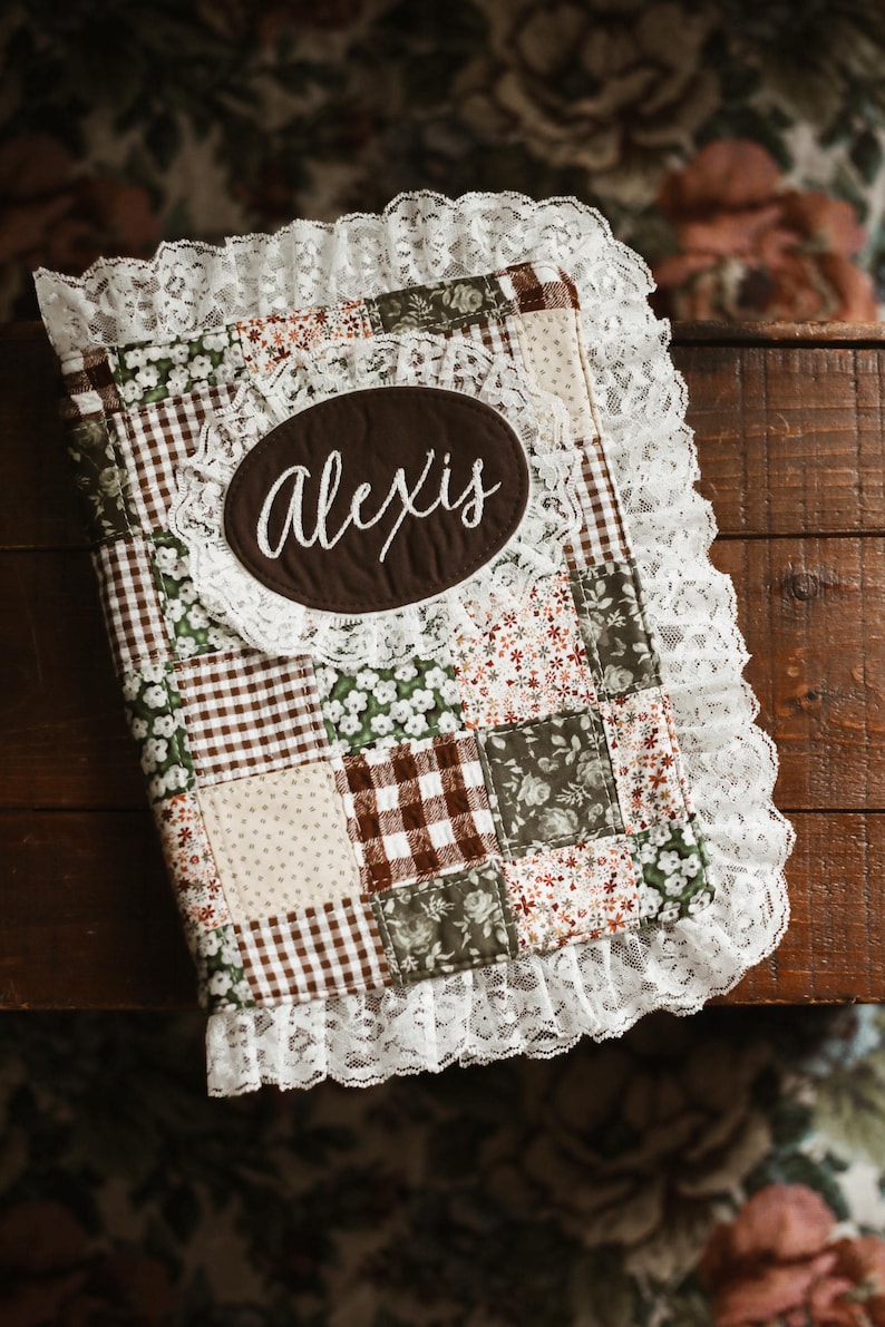 May include: A patchwork journal with a brown oval label embroidered with the name "Alexis" in white script. The journal features a variety of fabric patterns, including gingham, floral, and polka dots, and is trimmed with white lace.