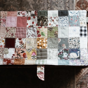 May include: A patchwork tote bag with a variety of floral and patterned fabric squares. The bag has light green handles and a matching fabric tab. The bag is sitting on a wooden surface.
