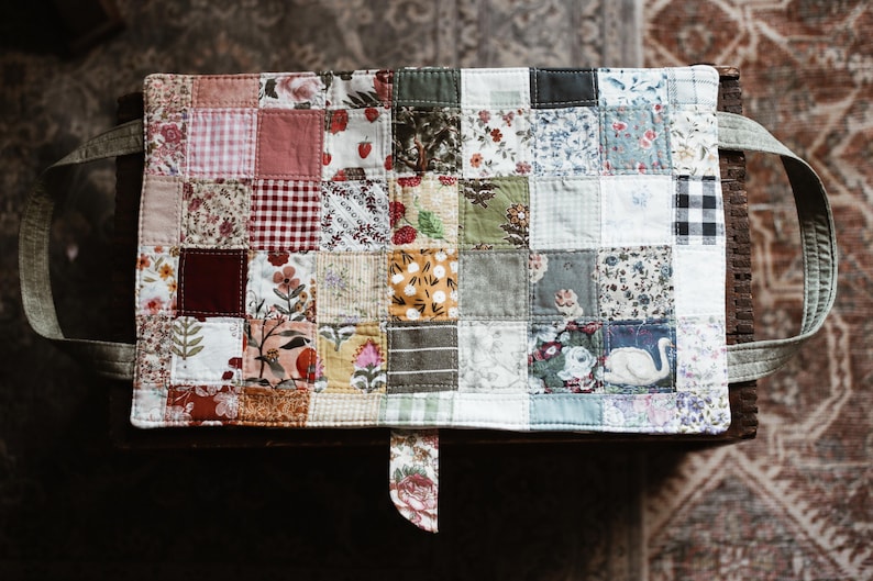 May include: A patchwork tote bag with a variety of floral and patterned fabric squares. The bag has light green handles and a matching fabric tab. The bag is sitting on a wooden surface.