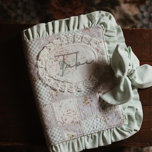 Könnte beinhalten: Ein stoffbezogenes Notizbuch mit einem Patchwork-Design aus Blumen- und Vichy-Karomustern in sanftem Lavendel und Weiß. Der Einband ist mit einem mintgrünen Rüschenrand, einer passenden seitlichen Schleife und einem ovalen Aufnäher verziert.