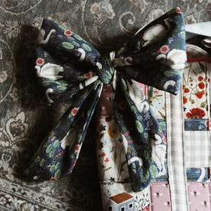 May include: A large, dark blue bow with a swan and floral pattern. The bow is tied and sits atop a patchwork quilt with various patterns, including houses, strawberries, and gingham. The background is a patterned gray fabric.