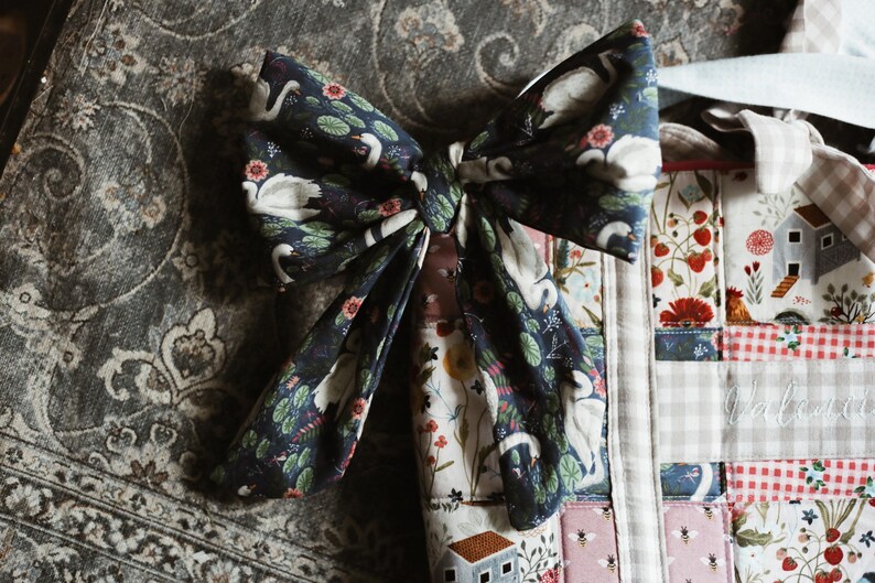 May include: A large, dark blue bow with a swan and floral pattern. The bow is tied and sits atop a patchwork quilt with various patterns, including houses, strawberries, and gingham. The background is a patterned gray fabric.