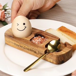 Peut inclure: Un ensemble de petit-déjeuner en bois sur une assiette blanche. Il comprend un œuf souriant, un compartiment à sel avec du sel rose, une cuillère dorée et deux tranches de pain grillé. Le texte "You're Cracking, Dad" est gravé.