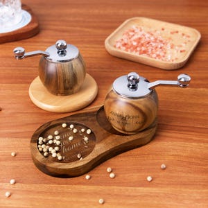 Puede incluir: Un juego de dos molinillos de sal y pimienta de madera con grabados personalizados. Los molinillos están sobre una bandeja de madera con un diseño de corazón y las palabras "some sweet home".