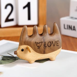Puede incluir: Una figurita de erizo de madera con un corazón y la palabra "LOVE" tallada en su espalda. La figurita está hecha de madera marrón claro y oscuro.