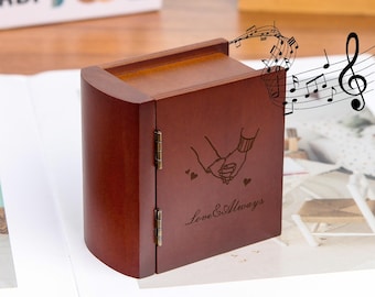 Puede incluir: Una caja de música de madera marrón oscuro con un diseño curvo. La parte frontal presenta una imagen grabada de dos manos tomadas de la mano, con las palabras "Love & Always" debajo. Se representan notas musicales encima de la caja.
