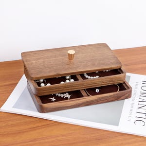 Könnte beinhalten: Eine rechteckige Schmuckschatulle aus Holz mit zwei Schubladen. Die Schatulle ist aus dunklem Holz gefertigt und hat einen goldfarbenen Knopf auf dem Deckel. Die Schubladen sind mit braunem Stoff ausgekleidet und enthalten Schmuck.