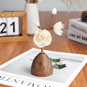May include: A wooden flower arrangement with a large cream-coloured flower and buds. The flower is mounted on a wooden base with the initials "DF" and a floral design. The arrangement sits on a white surface.