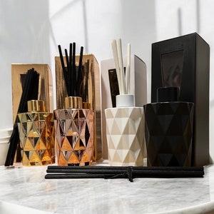 May include: Four decorative glass diffuser bottles with black, gold, rose gold, and white geometric designs. Each bottle has a set of reeds in it. The bottles are sitting on a white marble surface.