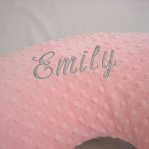 May include: A light pink nursing pillow with a dimpled texture, embroidered with the name "Emily" in grey script. The pillow is designed for comfort and support.