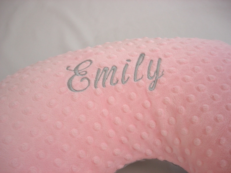 May include: A light pink nursing pillow with a dimpled texture, embroidered with the name "Emily" in grey script. The pillow is designed for comfort and support.