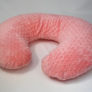 Op de afbeelding: Een roze, U-vormig voedingskussen met een textuur met stippen. Het kussen is ontworpen om moeders te ondersteunen tijdens het borstvoeden of voor baby's.
