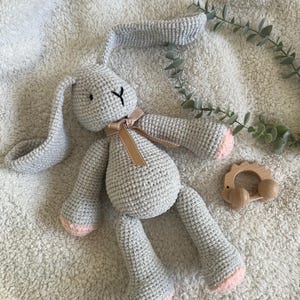 May include: A grey crocheted bunny toy with pink feet and a brown ribbon. A wooden teething ring sits beside the bunny on a soft, white blanket. The bunny has black embroidered eyes and a black 'Y' shaped mouth. The toy is suitable for a baby.