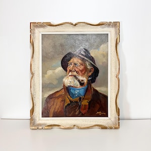 May include: Oil painting of an elderly man with a white beard, wearing a dark hat, brown jacket, and blue scarf, smoking a pipe. The artwork is framed in a decorative, off-white and gold frame. The background features a cloudy sky.