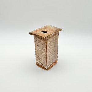 Puede incluir: Un jarrón rectangular de cerámica hecho a mano con un exterior marrón claro texturizado. La parte superior tiene una pequeña abertura circular y un tono marrón rojizo ligeramente más oscuro. La base tiene un borde blanco y marrón rojizo.
