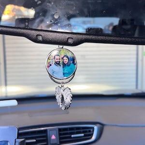 Op de afbeelding: Een zilveren autohanger met een ronde foto-inzet met een stel. De foto-inzet is bevestigd aan een zilveren hanger in de vorm van engelenvleugels.