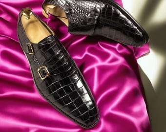 May include: A pair of black leather dress shoes with a crocodile skin texture. The shoes feature a double monk strap design with gold-colored buckles. The shoes are displayed on a vibrant pink satin fabric.