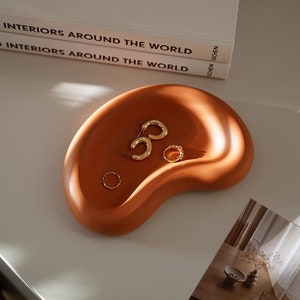 May include: A terracotta-colored ceramic dish in the shape of a kidney bean, with a set of gold hoop earrings and a gold ring inside. The dish is sitting on a white surface with a stack of books titled "100 Interiors Around the World" visible in the background.