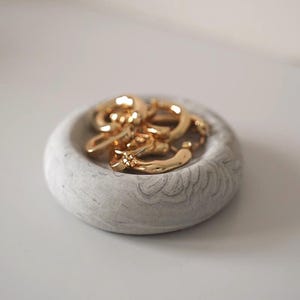 May include: A small, round, light grey concrete trinket dish with a subtle marbled texture and a carved floral pattern along the outer rim. Inside the dish sits a collection of shiny gold hoop earrings with a smooth, reflective metallic finish.