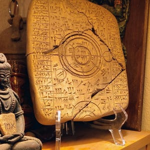 May include: A rectangular tan clay tablet featuring intricate cuneiform script and a central circular diagram with concentric rings. The tablet has a prominent diagonal crack running across the surface and is displayed on a clear plastic stand.