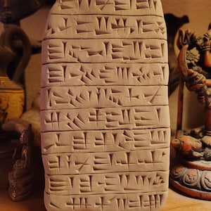May include: A beige clay tablet with cuneiform writing carved into its surface. The rectangular tablet stands upright, displaying the ancient script. Other decorative objects are visible in the background.