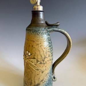 May include: A ceramic oil cruet with a mottled blue and tan glaze, featuring a raised dragonfly relief on the front. The vessel has a curved handle and a stainless steel pour spout with a hinged cap inserted into the narrow neck opening.