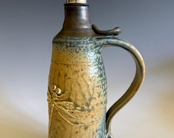 May include: A ceramic oil cruet with a mottled blue and tan glaze, featuring a raised dragonfly relief on the front. The vessel has a curved handle and a stainless steel pour spout with a hinged cap inserted into the narrow neck opening.