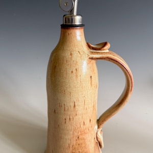May include: A ceramic oil cruet with a tan and brown speckled glaze, featuring a curved handle on the side and a stainless steel pour spout with a hinged lid inserted into the narrow neck of the bottle for controlled liquid dispensing.