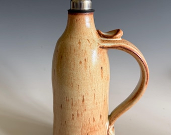 May include: A ceramic oil cruet with a tan and brown speckled glaze, featuring a curved handle on the side and a stainless steel pour spout with a hinged lid inserted into the narrow neck of the bottle for controlled liquid dispensing.