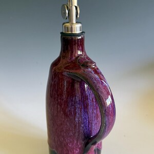 May include: A ceramic oil cruet featuring a glossy, mottled purple and magenta glaze over a solid black base. The vessel has a curved handle on the side and is topped with a silver-colored metal pour spout with a hinged cap for controlled dispensing.
