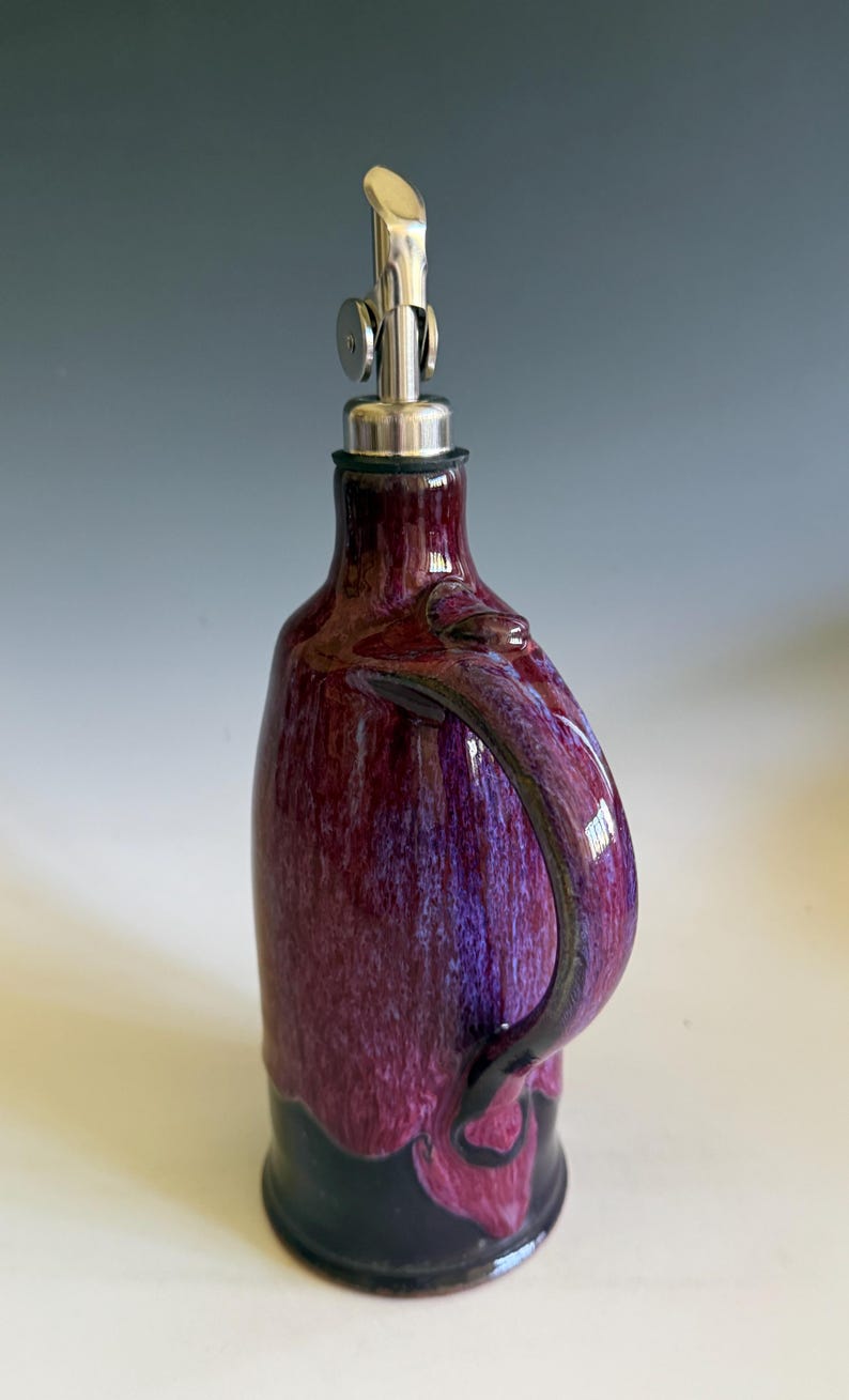 May include: A ceramic oil cruet featuring a glossy, mottled purple and magenta glaze over a solid black base. The vessel has a curved handle on the side and is topped with a silver-colored metal pour spout with a hinged cap for controlled dispensing.