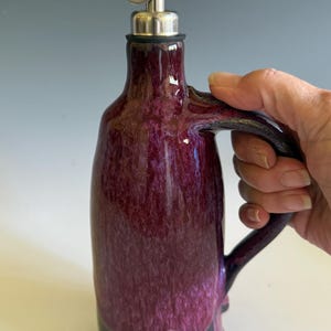 May include: A ceramic oil cruet featuring a glossy, mottled deep purple glaze on the upper body and a solid dark black base. The vessel includes a curved side handle and a stainless steel pour spout with a hinged metal cap at the top opening.