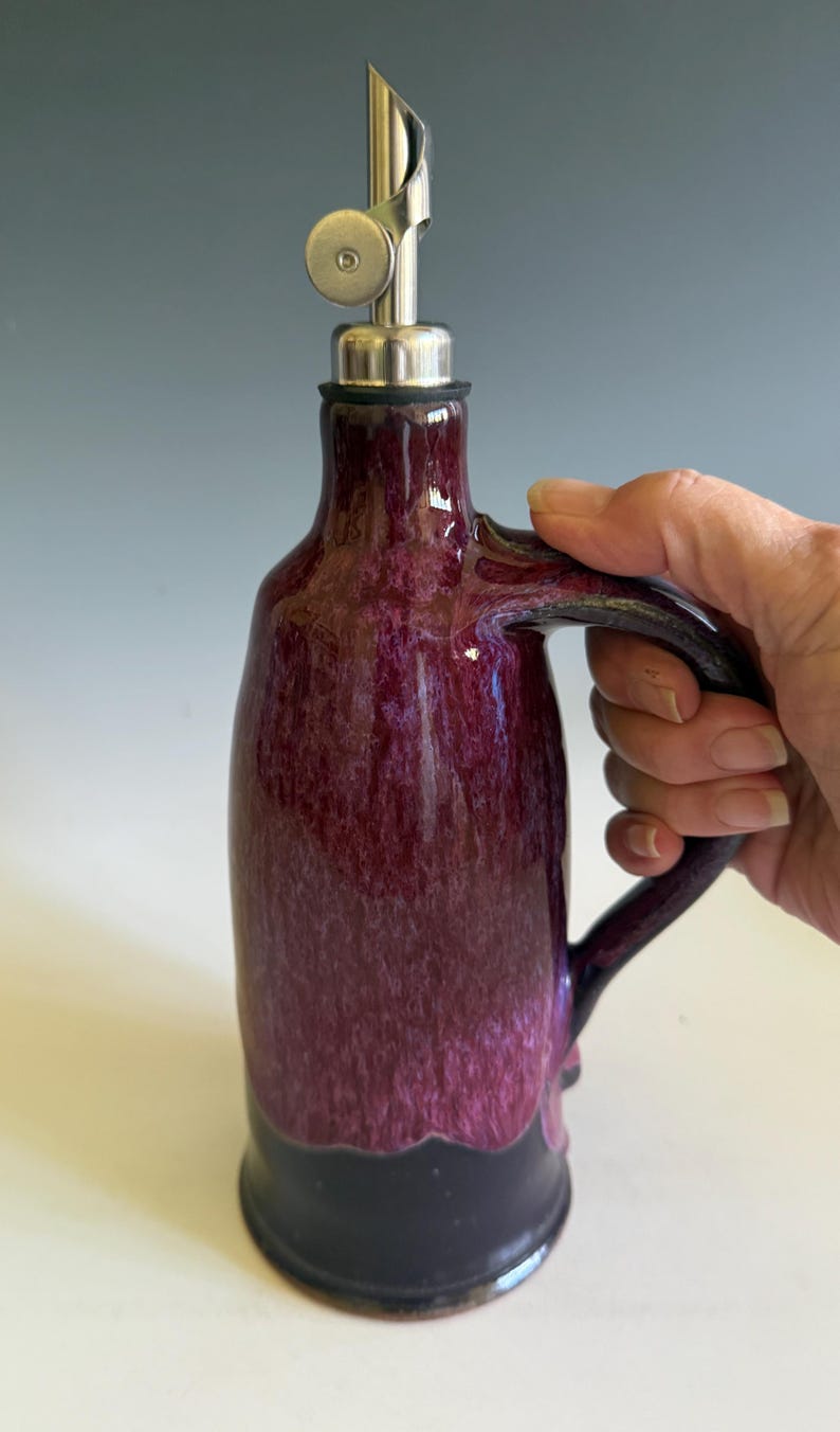 May include: A ceramic oil cruet featuring a glossy, mottled deep purple glaze on the upper body and a solid dark black base. The vessel includes a curved side handle and a stainless steel pour spout with a hinged metal cap at the top opening.