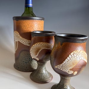 May include: A ceramic wine chiller and two matching goblets. The set features a brown and orange color scheme with a textured, crackled design. The goblets have a stem and a wide bowl. A green wine bottle is inside the chiller.