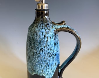 May include: A ceramic oil cruet featuring a tall cylindrical body with a handle. The base is solid dark brown, transitioning into a textured, dripped blue and white glaze on the upper portion. It is topped with a silver metal pour spout.