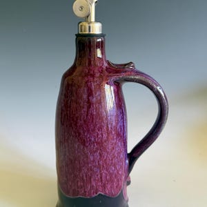 May include: A ceramic oil cruet featuring a glossy, mottled purple glaze on the upper body and a solid dark black base. The vessel includes a curved handle on the side and a stainless steel pourer spout with a hinged metal cap at the top.